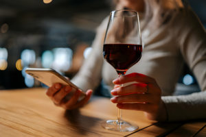 Personne tenant un verre de vin rouge et consultant son smartphone dans un bar.