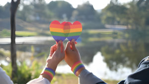 Deux personnes tenant ensemble un cœur aux couleurs du drapeau arc-en-ciel de la fierté, avec des bracelets assortis.