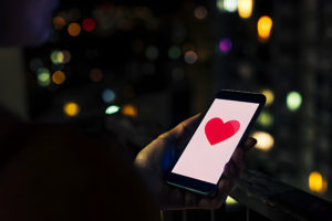 Une personne tenant un smartphone avec un cœur rouge sur l'écran, sur fond de lumières de ville floues.