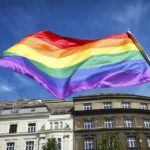 Drapeau arc-en-ciel de la fierté LGBTQ+ flottant fièrement devant un bâtiment en journée ensoleillée.
