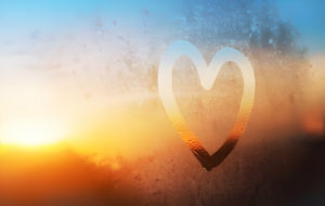 Cœur dessiné sur une fenêtre embuée avec un coucher de soleil en arrière-plan.