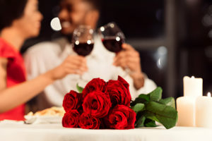 Un couple flou en arrière-plan portant un toast avec du vin rouge, un bouquet de roses rouges au premier plan.